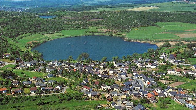 Eine malerische Landschaft mit einem ruhigen See und umgeben von grünen Feldern. Im Vordergrund ist ein kleines Dorf mit vielen Häusern zu sehen.