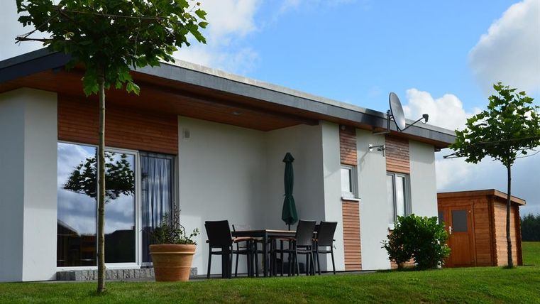 A modern house with a green garden. In front of the house, there is a table with chairs and a small garden shed.