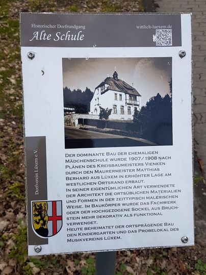 Ein historisches Schild, das die ehemalige Mädchenschule beschreibt. Das Gebäude ist von einer ländlichen Umgebung umgeben und zeigt architektonische Details.