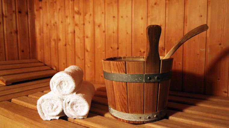 Ein hölzerner Saunaeimer mit drei zusammengerollten Handtüchern liegen auf einer Bank in einer Sauna.