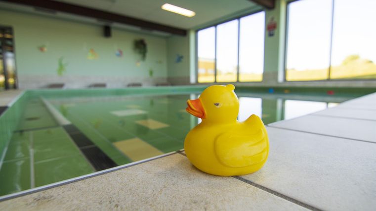 Een gele rubberen ligt aan de rand van een binnenzwembad. Het zwembad heeft een grote glazen gevel en heldere lijnen.