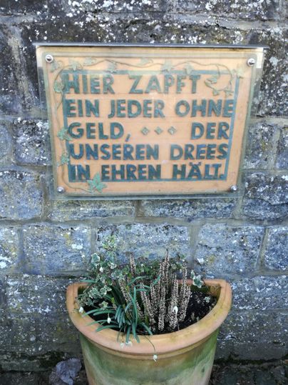 Ein altes Schild mit der Aufschrift: "Hier zapft ein jeder ohne Geld, der unseren Drees im Ehren hält." Im Hintergrund steht ein Blumentopf mit vertrockneten Pflanzen.