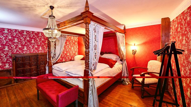 An elegant bedroom with a four-poster bed, surrounded by red wallpaper with floral patterns. The room contains a chair and a coat rack.