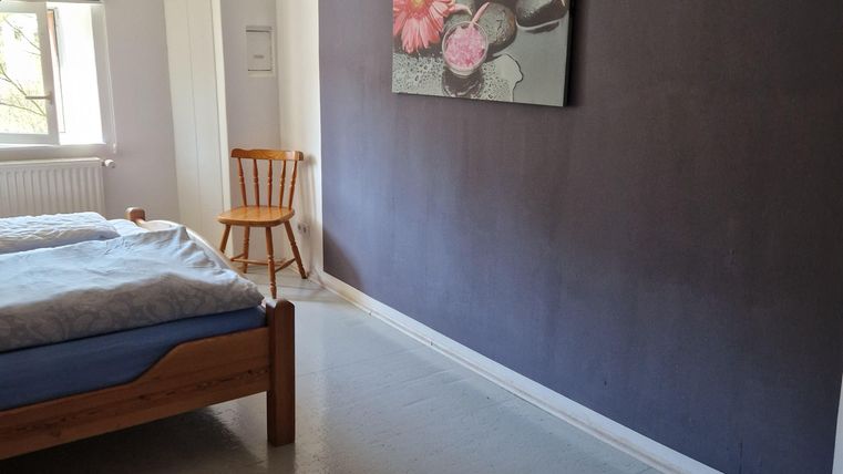 Een eenvoudige slaapkamer met een houten bed en een stoel. Aan de wand hangt een geschilderd beeld van bloemen.