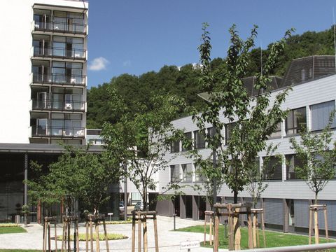 Moderne Gebäude mit klaren Linien und grünem Bereich. Bäume und Gemeinschaftsflächen schaffen eine einladende Atmosphäre.