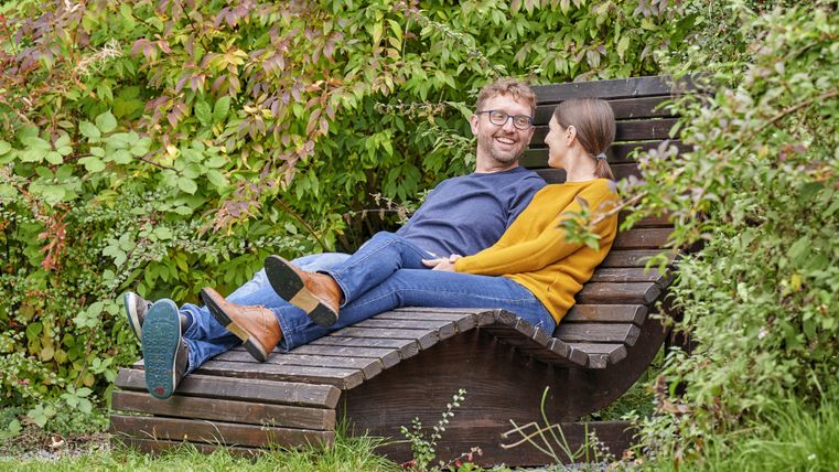 Ein Paar sitzt entspannt auf einer besonderen Holzliege inmitten von grünem Laub. Beide scheinen glücklich und genießen die Zeit miteinander.