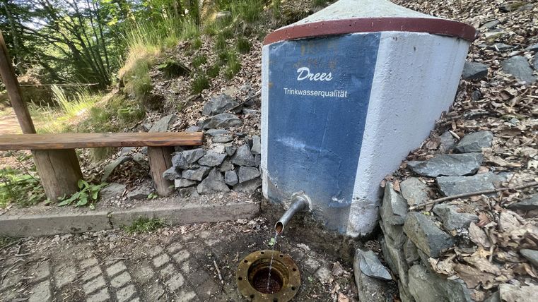 Een waterhuisje met een bron in de natuur. Naast het huisje staat een houten bank.