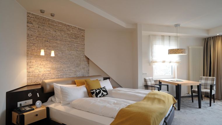 Ein modernes Schlafzimmer mit zwei Betten und hellen Farben. Es gibt einen Tisch mit Stühlen und große Fenster, die viel Licht hereinlassen.