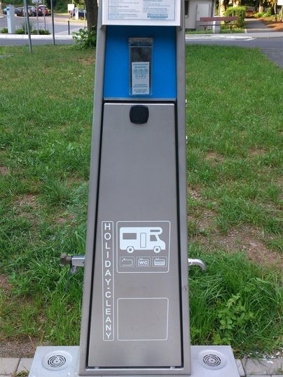 Ein moderner Geldautomat für Wohnmobile steht in einem Park. Er ist aus Metall und hat eine blaue Anzeige.