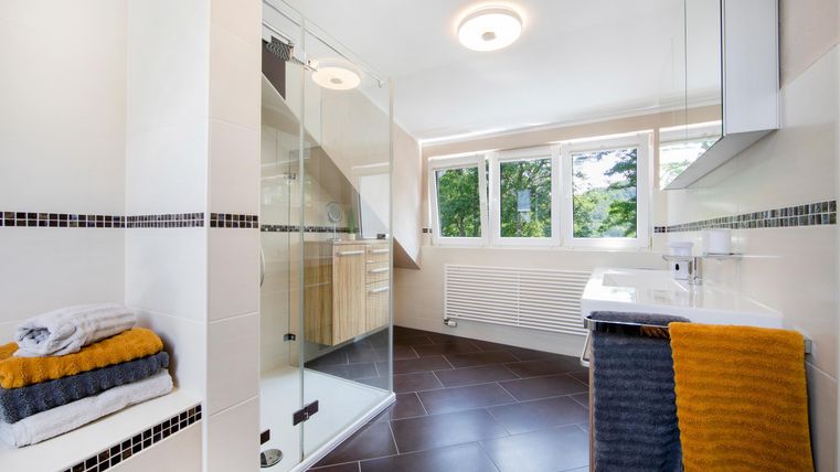 Ein modernes Badezimmer mit einer gläsernen Dusche und großen Fenstern. Handtücher in Blau und Gelb sind ordentlich gefaltet.