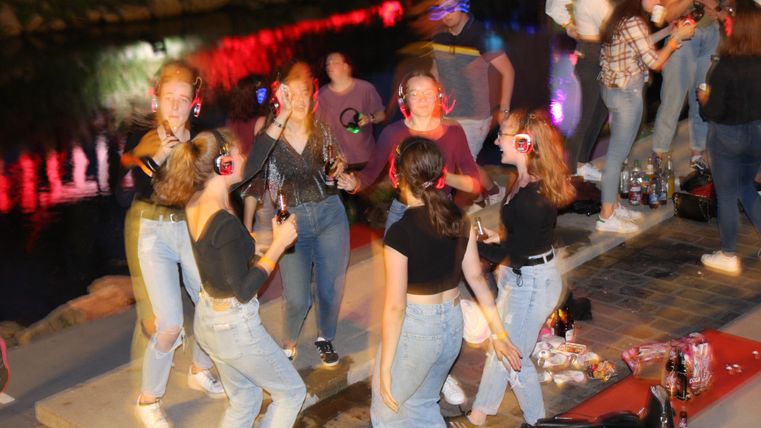 A group of young people is celebrating happily by a body of water. They are wearing headphones and dancing to music under the night lighting.