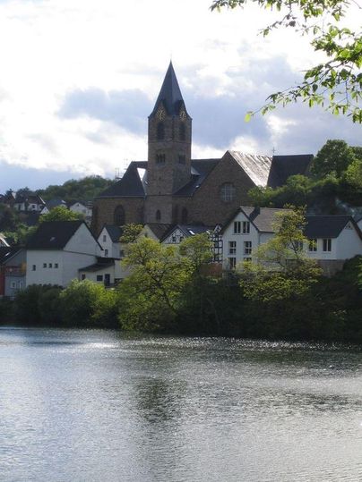 Een pittoreske kerk met een hoge toren staat aan de oever van een rustige meer. Omringd door groene bomen en kleurrijke gebouwen toont de scène een vredig landschap.