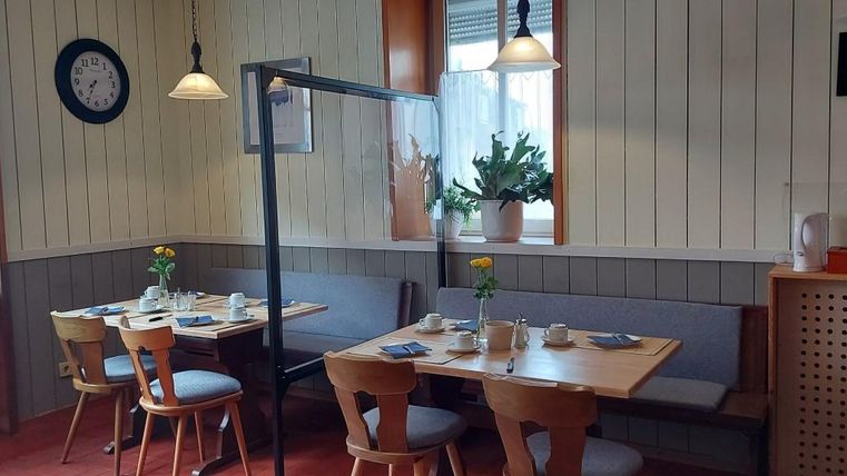 Ein gemütlicher Gastraum mit Holztischen und Stühlen. Auf dem Tisch sind gedeckte Plätze und Pflanzen in Töpfen zu sehen.