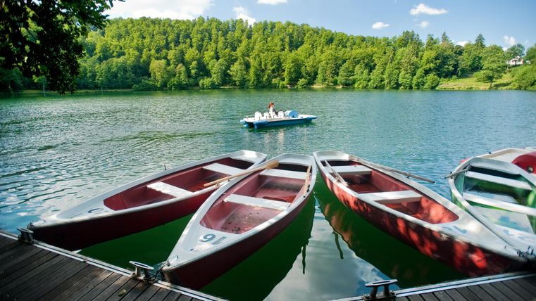 A peaceful lake landscape with boats at the shore. In the background, there are green trees and a few people in a boat.