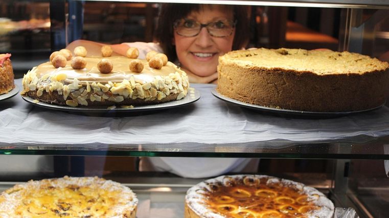 A friendly baker presents a selection of various cakes in a display case. The cakes are richly decorated and appetizing.