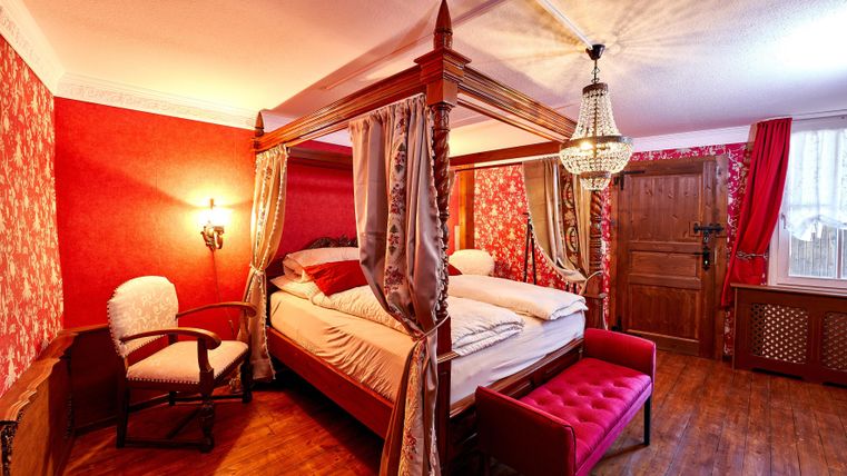 A cozy bedroom with a four-poster bed and red wallpaper. The room is stylishly furnished with an armchair and a crystal chandelier.