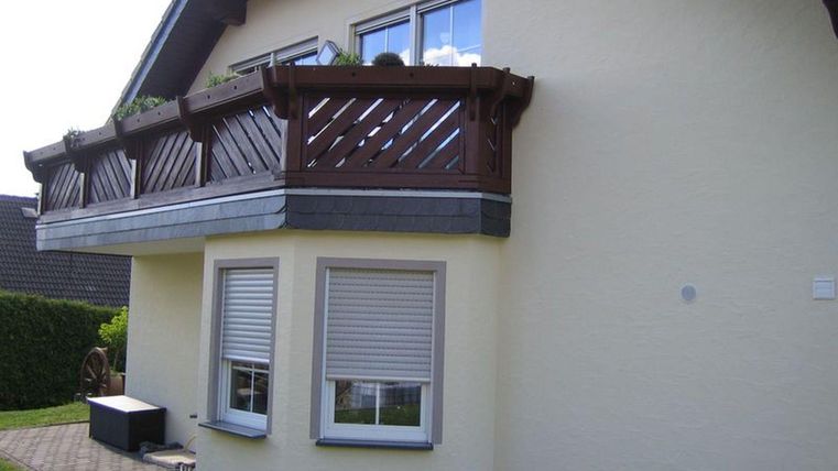 Een modern huis met een houten balkon. De gevel is geel en het perceel heeft een onderhouden tuin.