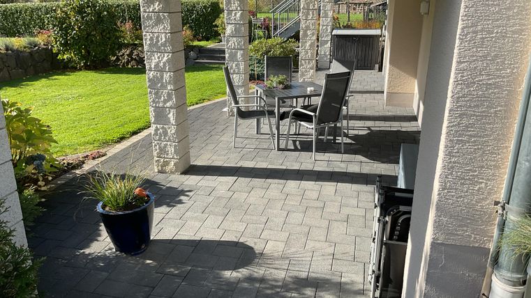 A cozy terrace with chairs and a table, surrounded by green grass and plants. The sun is shining on the paved floor.