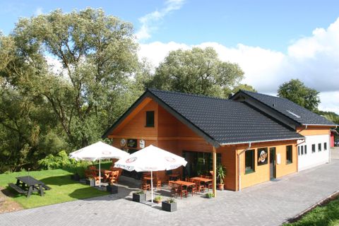 Holzgebäude mit Terrasse, Sonnenschirmen und Sitzgelegenheiten, umgeben von Bäumen und blauem Himmel.