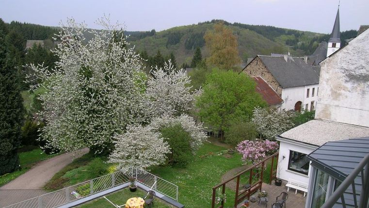 Eine malerische Landschaft mit blühenden Bäumen und sanften Hügeln im Hintergrund. Im Vordergrund sieht man einen Garten mit einer Terrasse und einem kleinen Tisch.