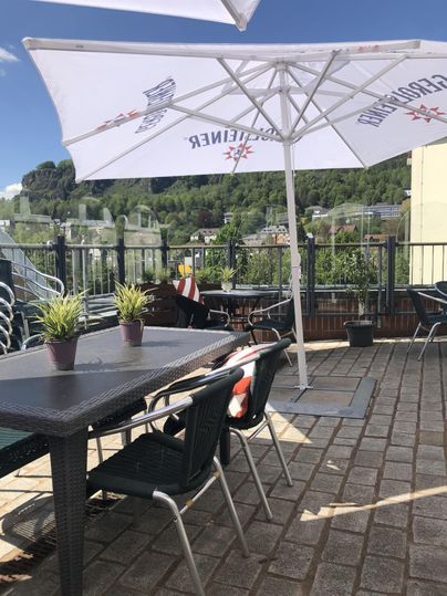 Terrace of the bistro with tables, chairs, and umbrellas. In the background, green hills and a blue sky.