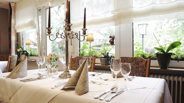 Ein elegant gedeckter Tisch mit Kerzen und Gläsern. Im Hintergrund sind Pflanzen und Fenster zu sehen, die für eine gemütliche Atmosphäre sorgen.