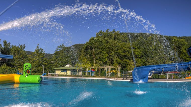 Een zwembad met helder blauw water en waterattracties. Op de achtergrond zijn groene bomen en een glijbaan te zien.