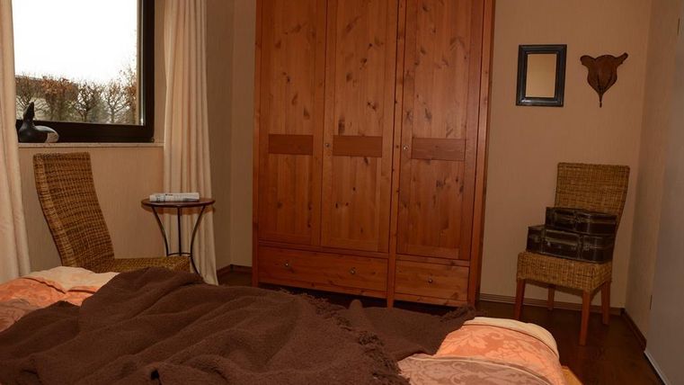 A cozy bedroom with a wooden wardrobe and two chairs. The bed is adorned with a brown blanket and colorful pillows.