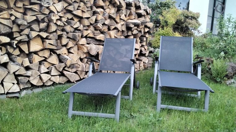 Zwei Liegen auf einer grünen Wiese, neben einem Holzstapel. Im Hintergrund ist ein Gebäude mit großen Fenstern zu sehen.