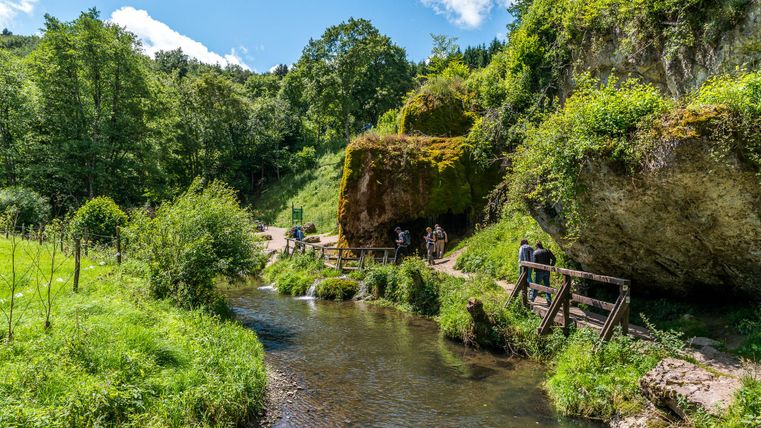 Ein Bach fließt durch eine grüne Landschaft mit Bäumen und Felsen. Menschen spazieren auf einem Pfad entlang des Wassers.