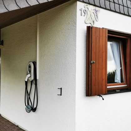 Een muur met een laadstation voor elektrische auto's en een open raam. De gevel is wit met een houten raam.