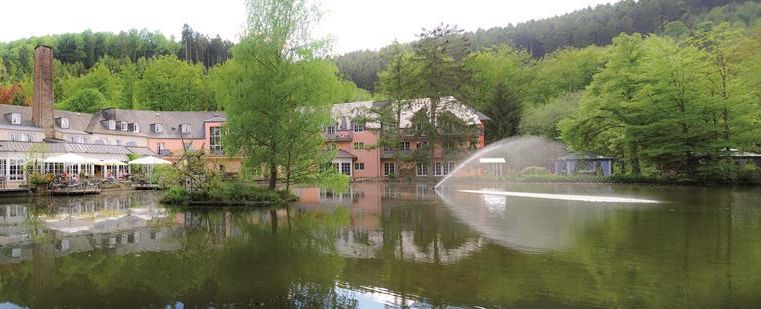 A picturesque lake with green trees and a modern building in the background. A water feature gently sprays in the middle of the lake.