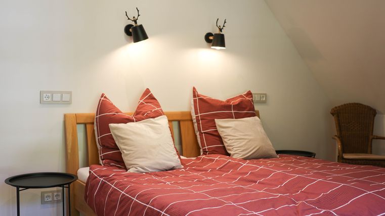 A cozy bedroom with a double bed and red pillows. Stylish lamps hang on the wall, and a wicker chair stands next to the bed.