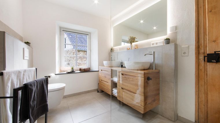 Ein modernes Badezimmer mit hellem Design und Holzdetails. Es verfügt über zwei Waschbecken, einen großen Spiegel und ein Fenster mit Blick nach außen.