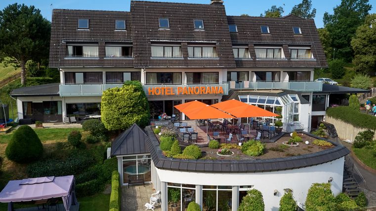 Een modern hotel met het opschrift "Hotel Panorama". Het heeft een terras met oranje parasols en is omringd door verzorgde tuinen.