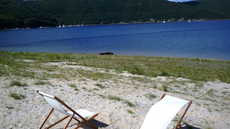 Zwei Liegestühle stehen am Strand neben einem ruhigen Gewässer. Im Hintergrund sind sanfte Hügel und ein klarer Himmel zu sehen.