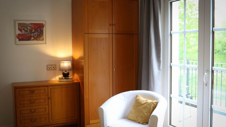 A cozy room with a white armchair and a wooden box. The window lets in plenty of daylight and offers a view of the greenery.