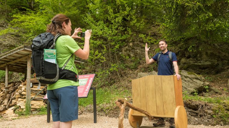 Eine Frau fotografiert einen Mann, der hinter einem hölzernen Wagen steht, auf einem Wanderweg im Wald.