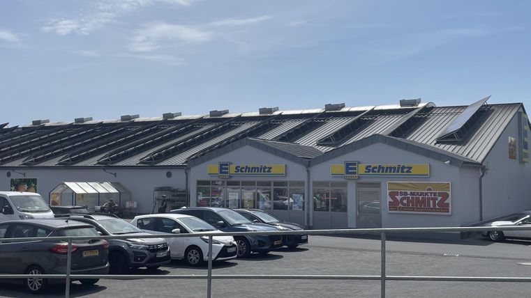 A commercial building named "Schmitz" with a modern roof. In front of the building, several cars are parked on a large parking lot.