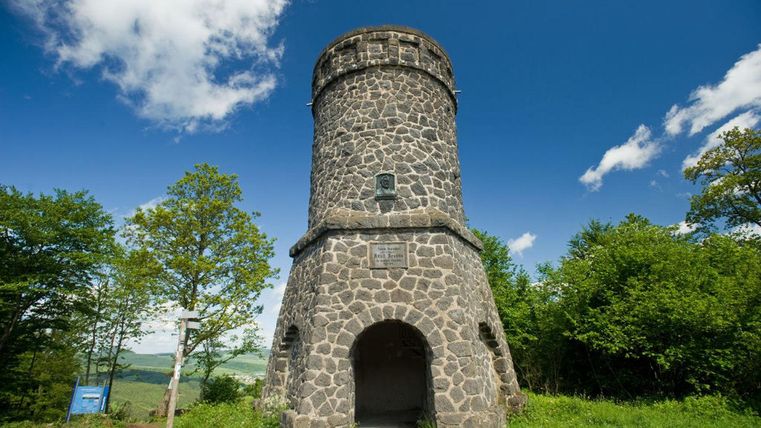 A historic stone tower stands surrounded by lush greenery and blue sky. The sun is shining, and the clouds are partially visible.