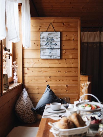 A cozy room with wooden walls and a table adorned with fresh bread and a fruit bowl. Cushions in various colors invite relaxation.