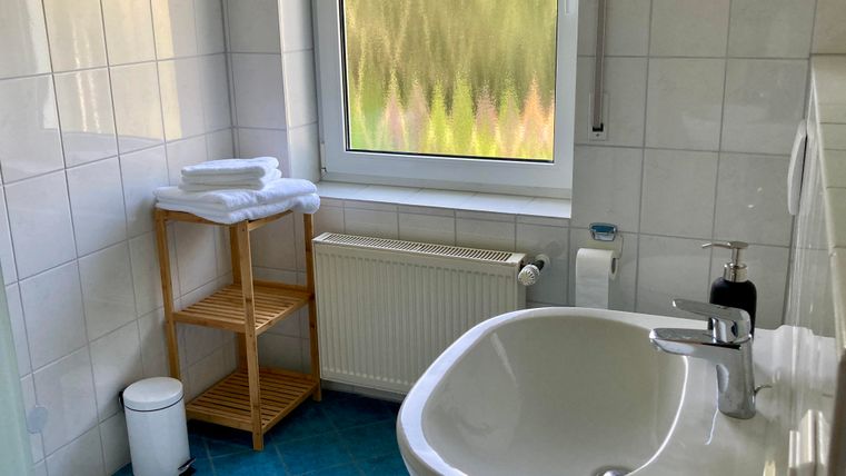 Ein modernes Badezimmer mit einem Waschbecken und einem Fenster. Es gibt auf einem Regal frische Handtücher und einen kleinen Abfalleimer.