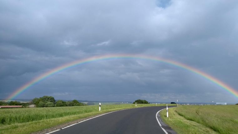 A winding road under a colorful rainbow. The sky is overcast, with green meadows on both sides.