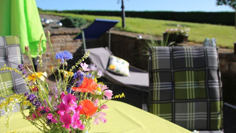 Ein schöner Tisch mit bunten Blumen in einer Vase. Im Hintergrund sind bequeme Stühle und ein sonniger Garten zu sehen.