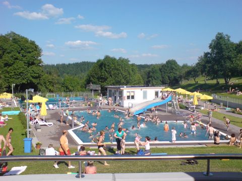 Ein belebtes Freibad mit vielen Badegästen und einem großen Pool. Sonnenschirme und Liegen umgeben den Bereich, darunter grünes Gras und Bäume.