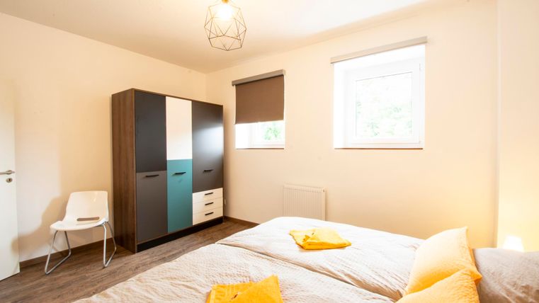 Ein helles Schlafzimmer mit zwei Betten und einem modernen Kleiderschrank. Auf den Betten liegen orangefarbene Kissen und die Wände sind neutral gehalten.