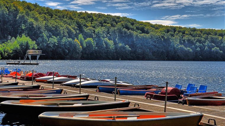 Ein ruhiger See mit Booten am Ufer. Im Hintergrund sind grüne Bäume und ein blauer Himmel zu sehen.