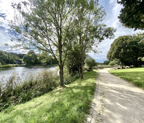 Ein sonniger Park mit einem geschwungenen Weg entlang eines Sees, umgeben von B&auml;umen und gr&uuml;nen Wiesen., &copy; Touristik GmbH Gerolsteiner Land
