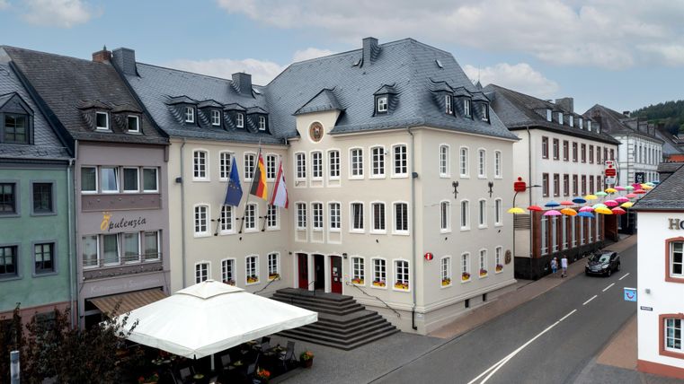 A historic building with multiple flags in front of the facade. On the street, colorful bunting and a restaurant with a sun umbrella can be seen.
