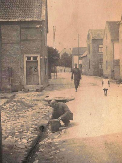 Een historisch straatbeeld met stenen en oude huizen. Een man is op de grond aan het werk, terwijl een persoon en een kind voorbijlopen.